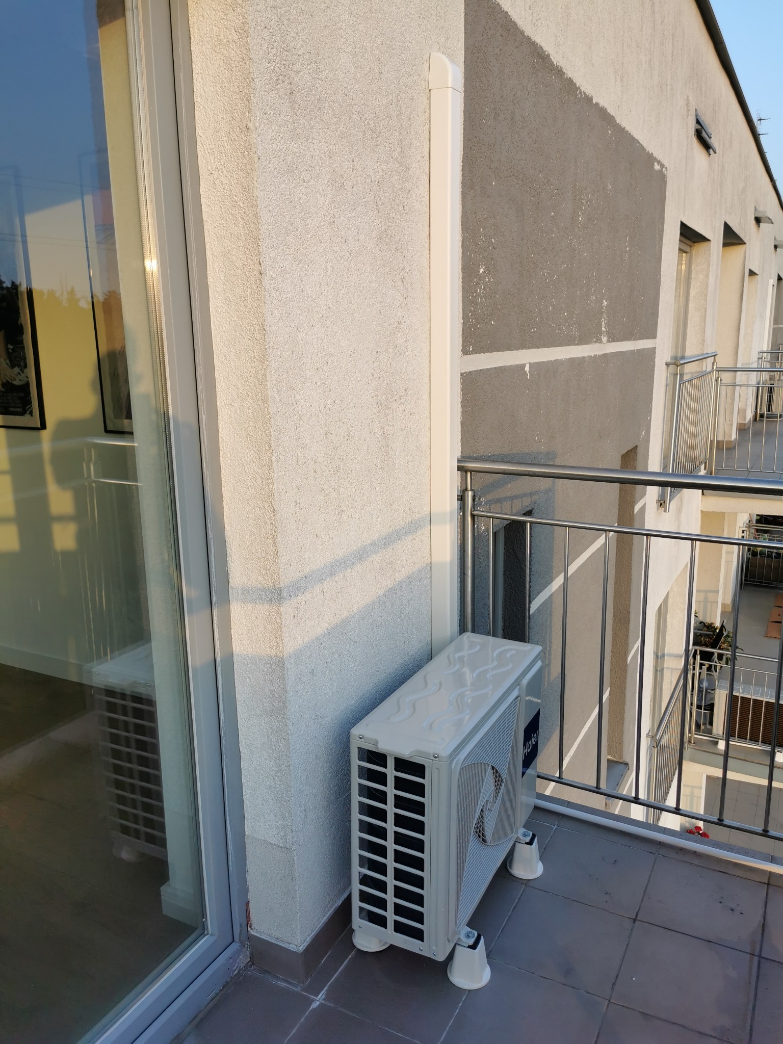 Biała jednostka zewnętrzna klimatyzacji, marki Haier, zamontowana na balkonie nowoczesnego apartamentowca z widocznymi metalowymi barierkami i szarą elewacją, oświetlona popołudniowym słońcem.