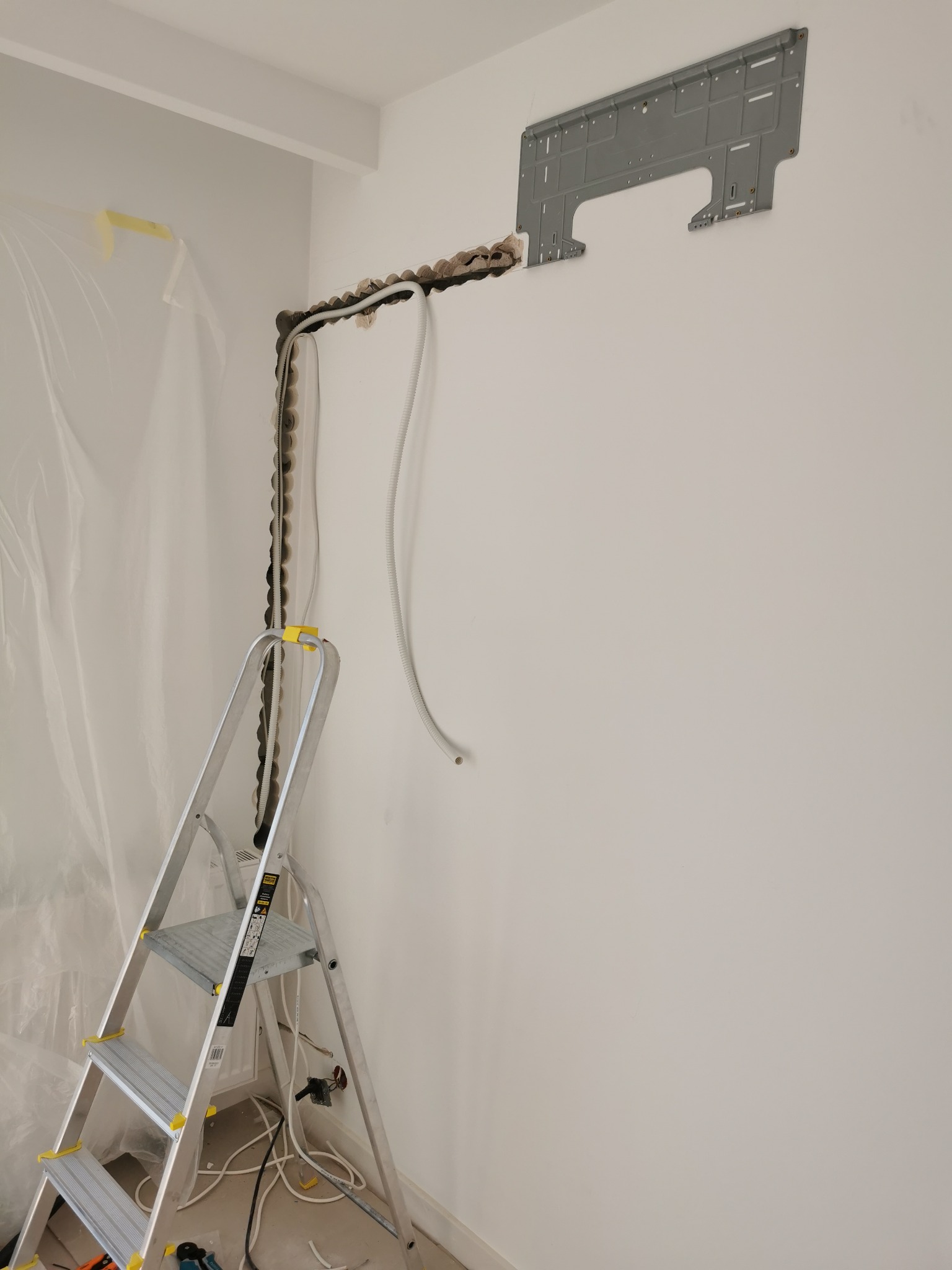 Przygotowanie do montażu jednostki wewnętrznej: wycięty otwór w ścianie, zamocowana listwa montażowa, widoczne przewody i drabina aluminiowa z żółtymi elementami zabezpieczającymi, folia ochronna...