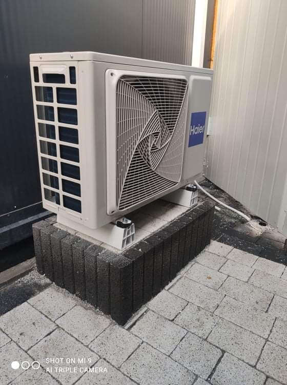 Zewnętrzna jednostka klimatyzacji Haier zamontowana na podwyższeniu z szarej kostki brukowej, widoczny kabel zasilający i ślady montażu na kostce.