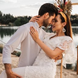 Maggieem studio - Zakochana para w objęciach na tle jeziora i dekoracji z suszonych kwiatów; kobieta w koronkowej sukience i opasce na włosach, mężczyzna w białej koszuli.