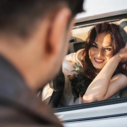 Maggieem studio - Uśmiechnięta kobieta z bukietem opiera się o okno samochodu, spoglądając na mężczyznę poza kadrem.