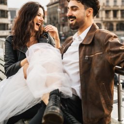 Maggieem studio - Uśmiechnięta para siedzi na ławce w miejskim otoczeniu: kobieta w czarnej kurtce i tiulowej spódnicy, mężczyzna w skórzanej kurtce, oboje patrzą na siebie z radością.