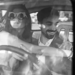 Czarno-białe zdjęcie pary siedzącej w samochodzie, kobieta w dużych okularach przeciwsłonecznych, mężczyzna uśmiechnięty, widoczny przez szybę z odbiciami.