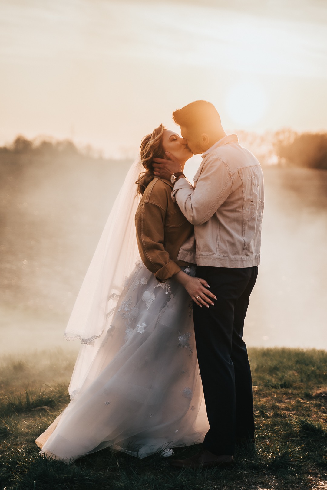 Para młoda całuje się na tle jeziora o zachodzie słońca, panna młoda w sukni z aplikacjami kwiatowymi i dżinsowej kurtce, pan młody w jasnej kurtce dżinsowej z haftem na plecach.