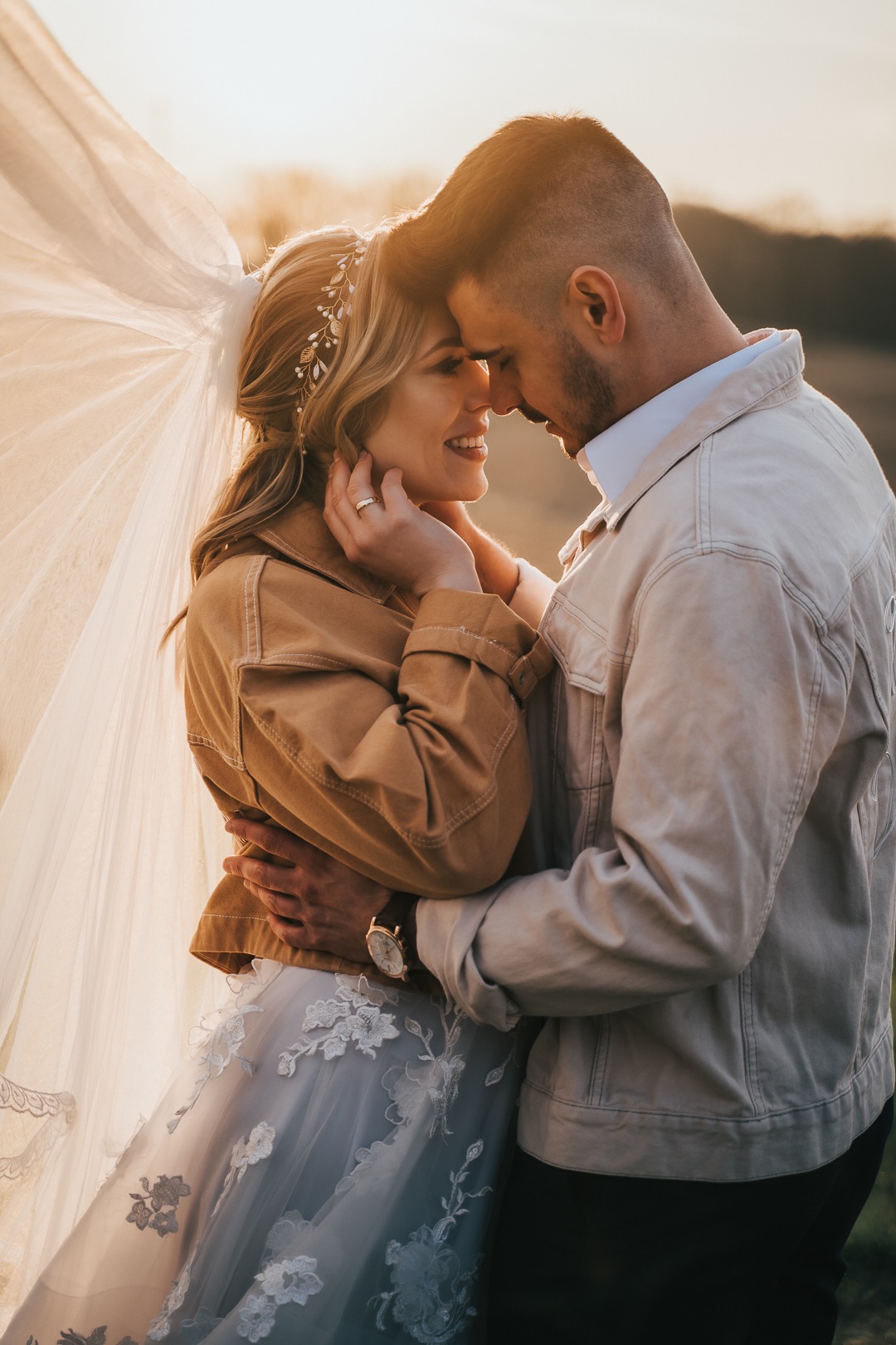 Uśmiechnięta panna młoda w dżinsowej kurtce i tiulowej sukni z koronkowymi aplikacjami, obejmowana przez pana młodego w dżinsowej kurtce, podczas sesji w plenerze o zachodzie słońca.