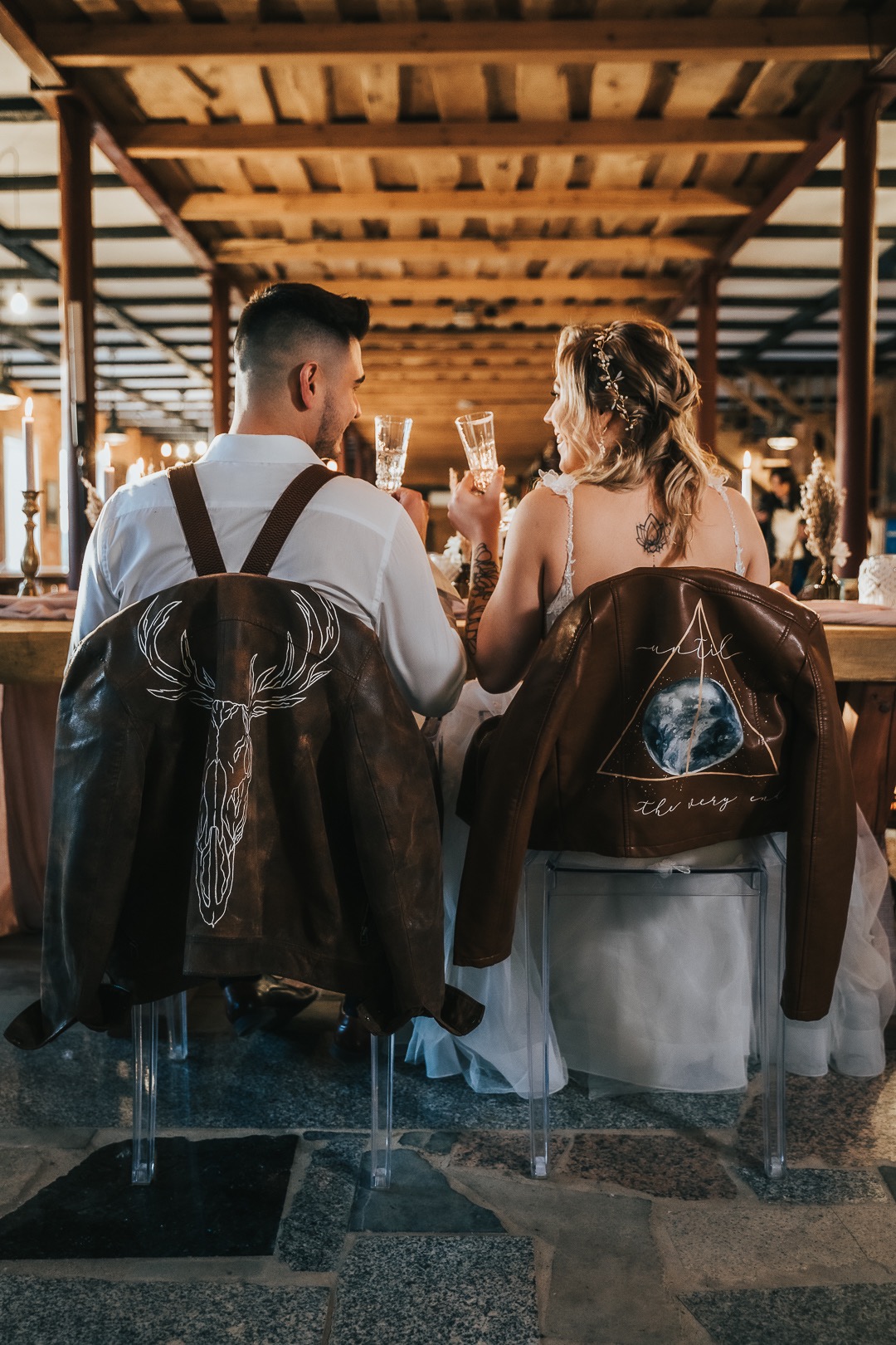 Para młoda siedzi przy stole, trzymając kieliszki z szampanem; na oparciach krzeseł zawieszone skórzane kurtki z ręcznie malowanymi wzorami: poroże jelenia i motyw kosmiczny.