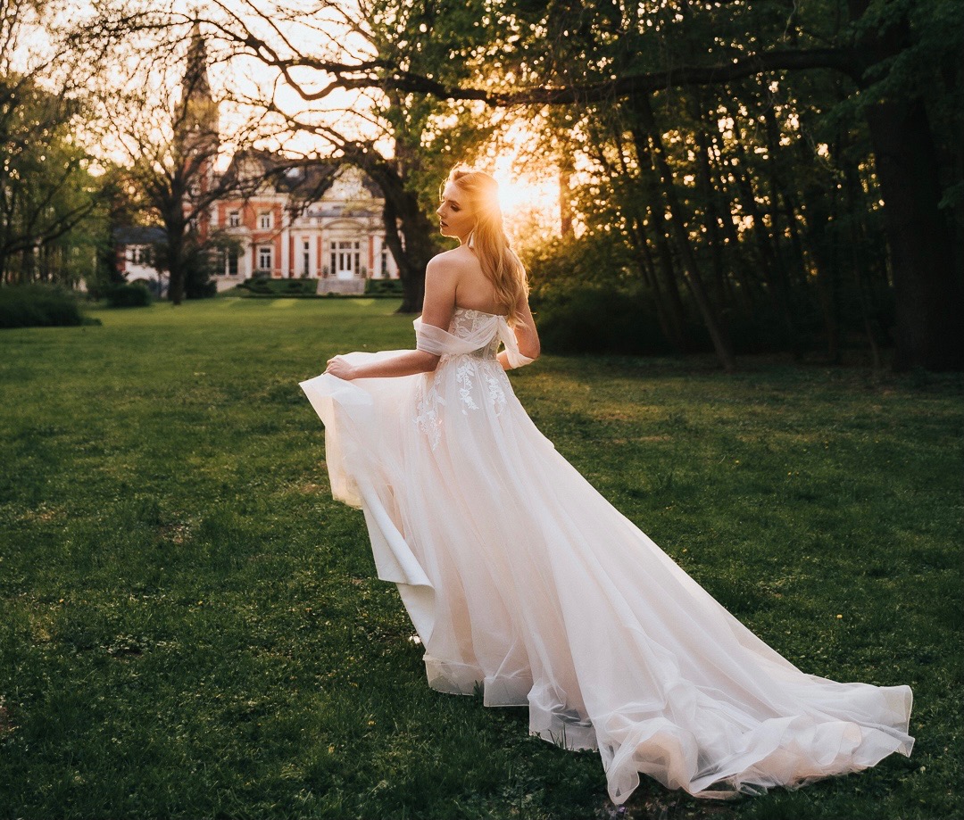 Panna młoda w zwiewnej sukni ślubnej z długim trenem, odwrócona plecami do kamery, na tle parku i zabytkowego pałacu w promieniach zachodzącego słońca.