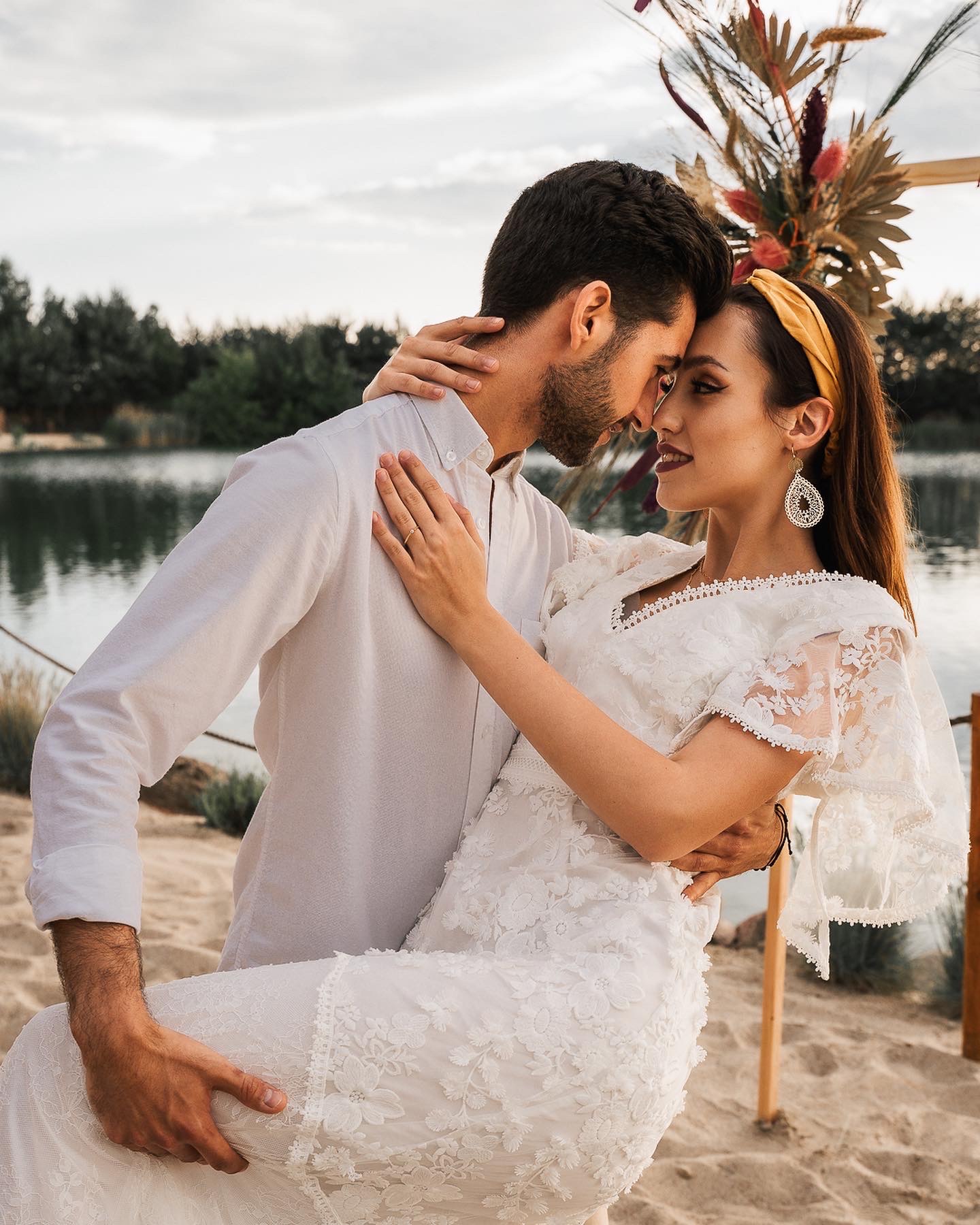 Zakochana para w objęciach na tle jeziora i dekoracji z suszonych kwiatów; kobieta w koronkowej sukience i opasce na włosach, mężczyzna w białej koszuli.