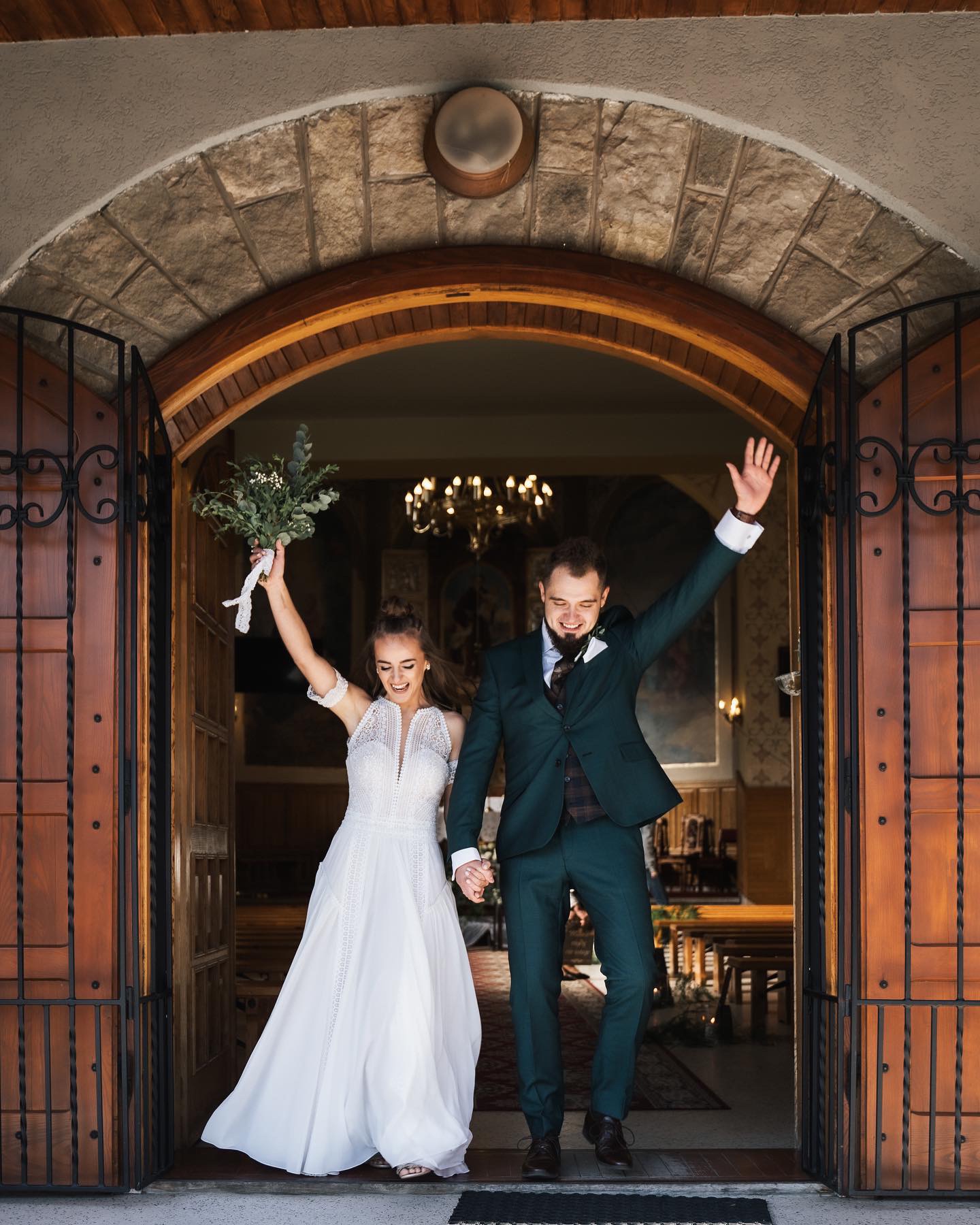 Para młoda, trzymając się za ręce i unosząc ręce w geście radości, wychodzi z kościoła przez kamienny portal z ozdobnymi, kutymi drzwiami. Panna młoda w białej sukni z bukietem, pan młody...