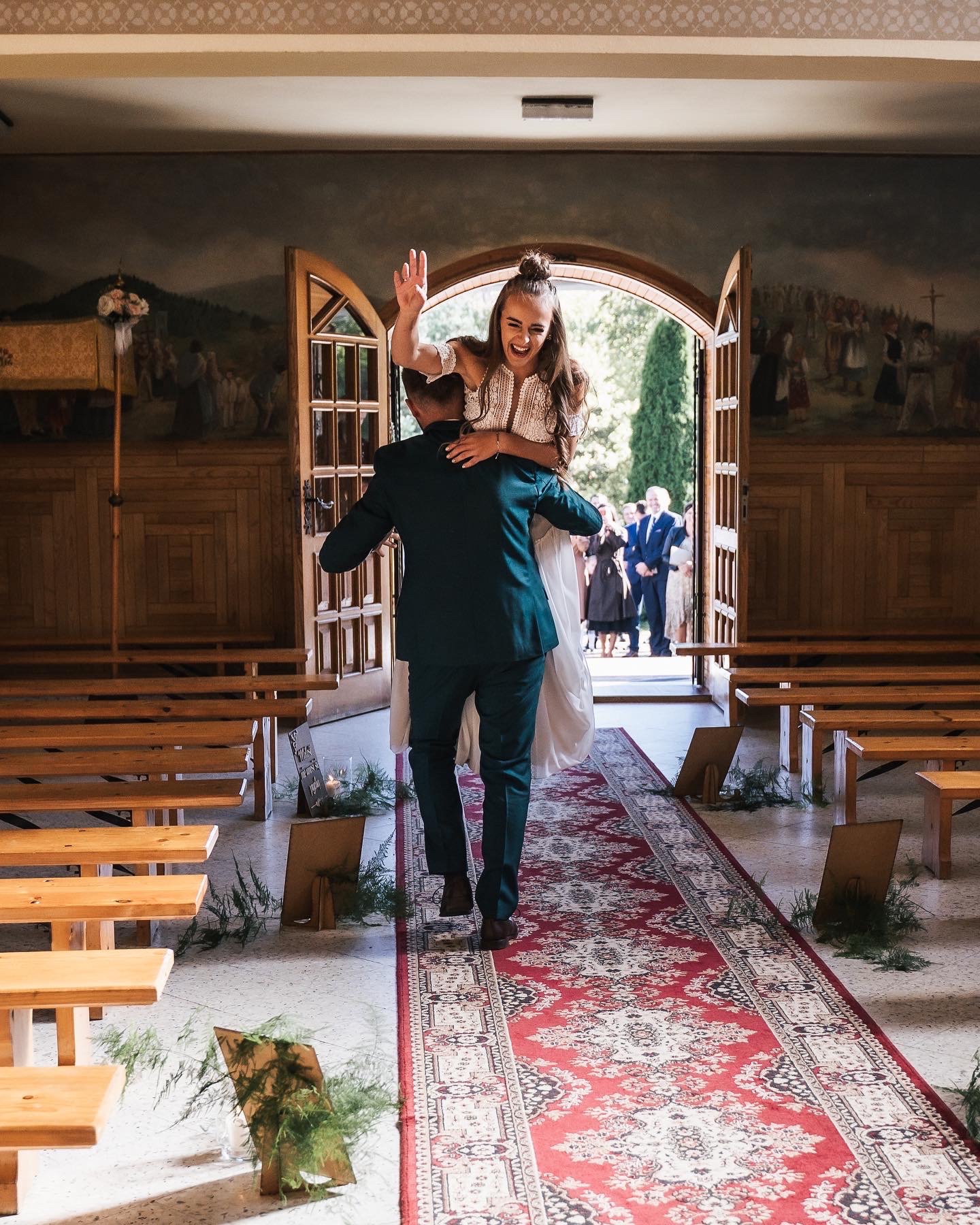 Pan młody w ciemnozielonym garniturze niesie uśmiechniętą pannę młodą na rękach, wychodząc z kościoła udekorowanego drewnianymi ławkami i freskami.