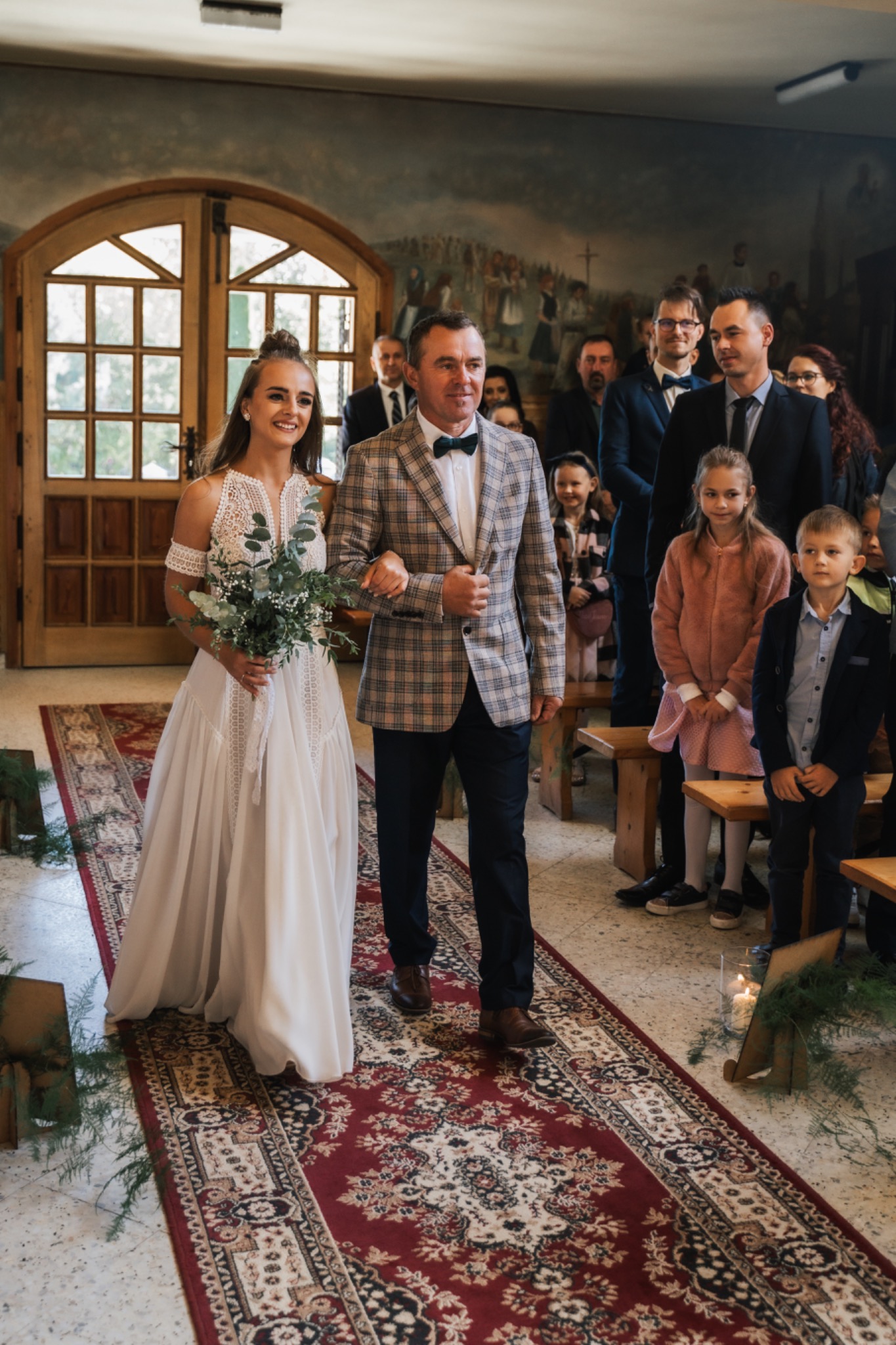 Panna młoda w koronkowej sukni i mężczyzna w kraciastej marynarce idą czerwonym dywanem w kościele, za nimi zgromadzeni goście.