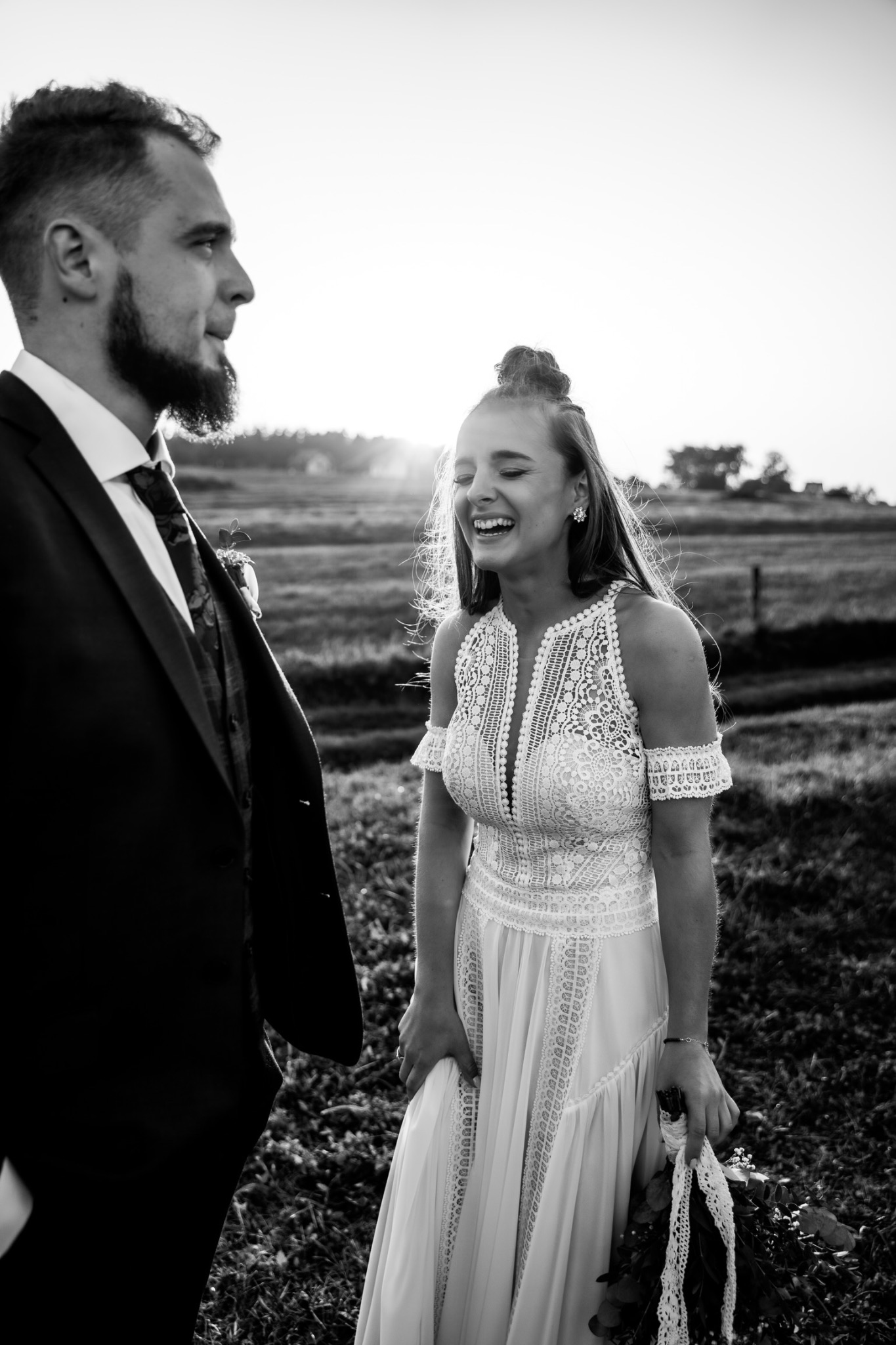 Czarno-białe zdjęcie uśmiechniętej panny młodej w koronkowej sukni, stojącej obok pana młodego w garniturze na tle łąki o zachodzie słońca.