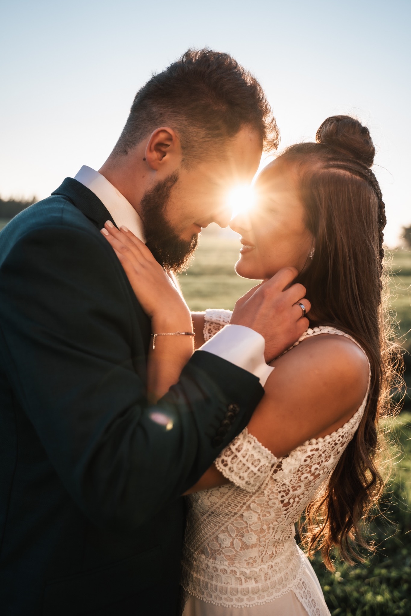 Para młoda w objęciach, promienie zachodzącego słońca rozświetlają twarz kobiety, widoczny pierścionek na palcu mężczyzny, ona w koronkowej sukni, on w ciemnozielonym garniturze.