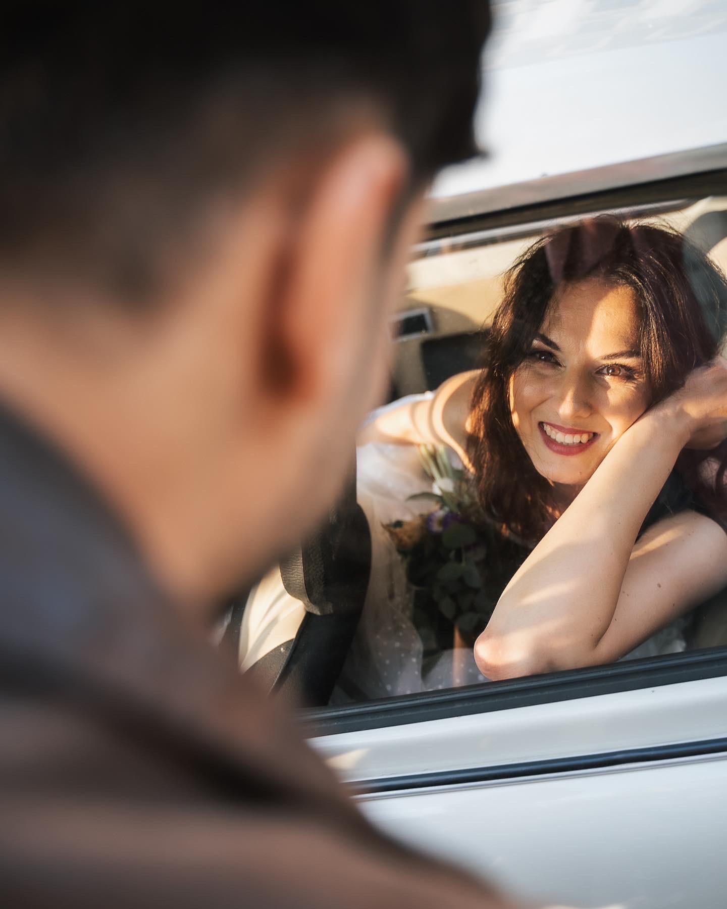 Uśmiechnięta kobieta z bukietem opiera się o okno samochodu, spoglądając na mężczyznę poza kadrem.