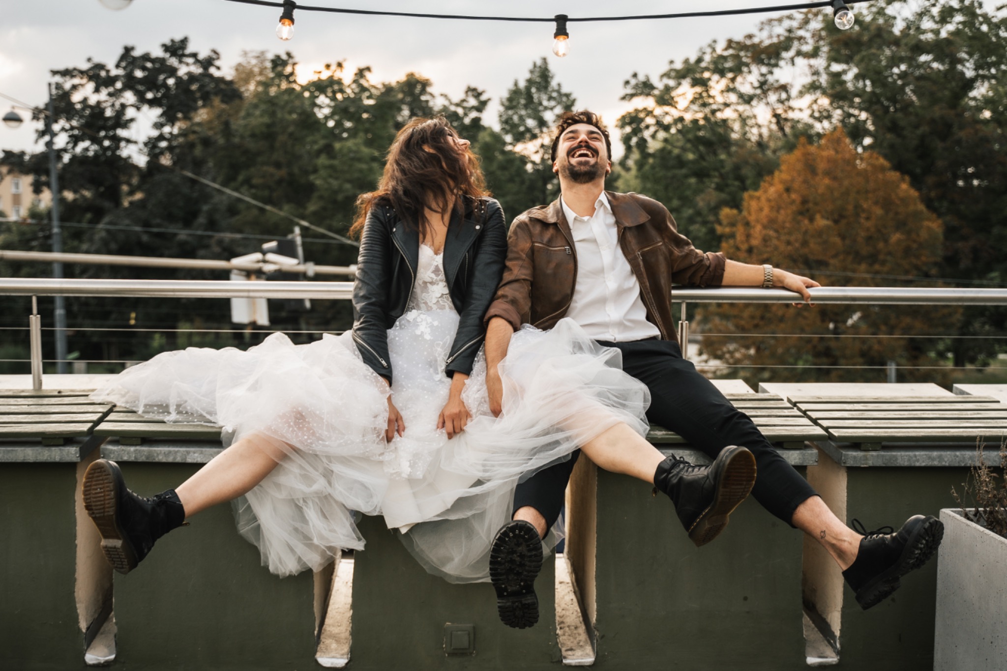 Uśmiechnięta para młoda siedzi na murku z nogami zwisającymi w dół; panna młoda w sukni ślubnej i czarnej skórzanej kurtce, pan młody w brązowej kurtce i czarnych spodniach, oboje w czarnych butach...