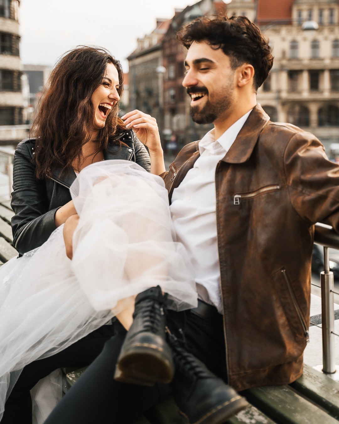 Uśmiechnięta para siedzi na ławce w miejskim otoczeniu: kobieta w czarnej kurtce i tiulowej spódnicy, mężczyzna w skórzanej kurtce, oboje patrzą na siebie z radością.