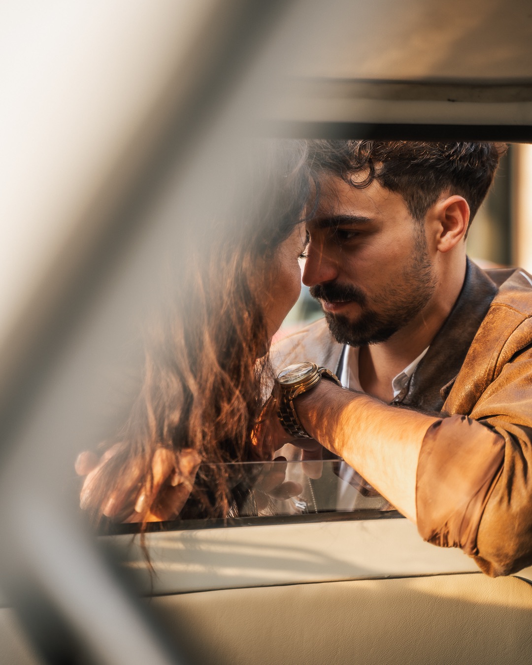 Para zakochanych w samochodzie, zbliżenie twarzy, złoty zegarek na nadgarstku mężczyzny, skórzana kurtka, kręcone włosy kobiety, ciepłe światło.