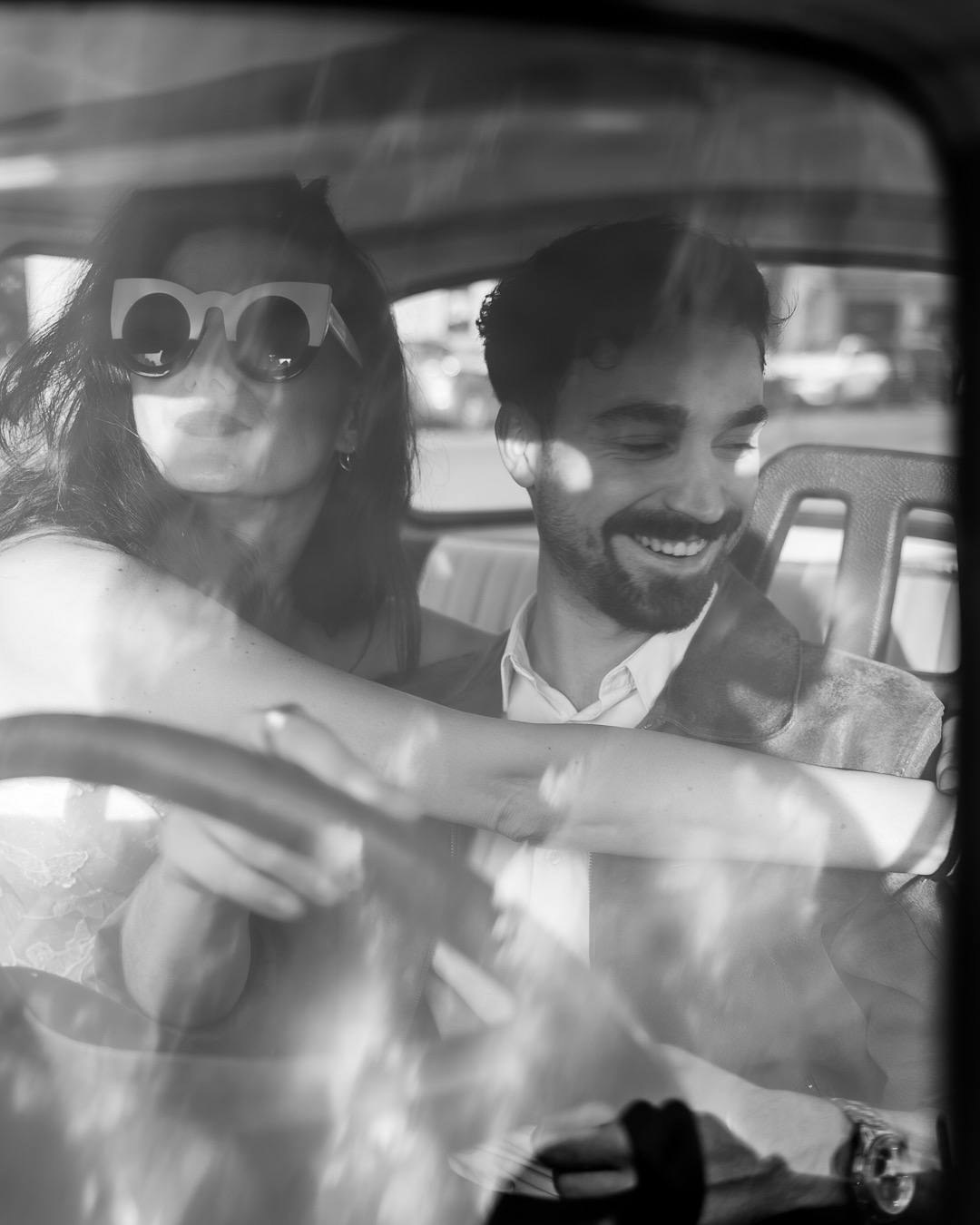 Czarno-białe zdjęcie pary siedzącej w samochodzie, kobieta w dużych okularach przeciwsłonecznych, mężczyzna uśmiechnięty, widoczny przez szybę z odbiciami.