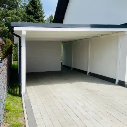 Nowoczesna, biała wiata garażowa z płaskim dachem w Oleśnicy, przylegająca do domu, z kostką brukową na podjeździe i metalową siatką ogrodzeniową po lewej stronie.