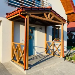 Zadaszone wejście do domu z drewnianą konstrukcją, ozdobną balustradą krzyżową i niebieskimi drzwiami z numerem 62, udekorowane wieńcem.
