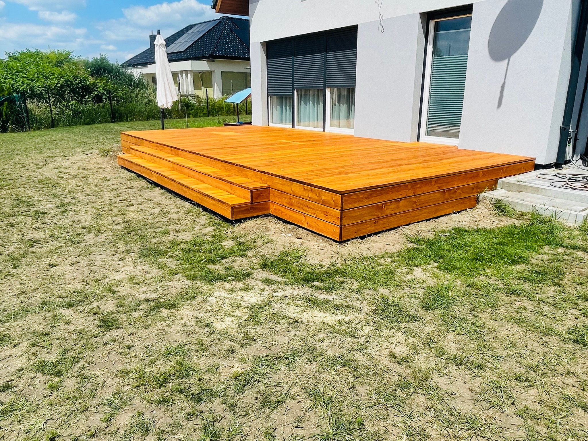 Nowoczesny, drewniany taras z trzema stopniami, przylegający do jasnego domu z roletami zewnętrznymi, otoczony trawnikiem z widocznymi przeschnięciami.