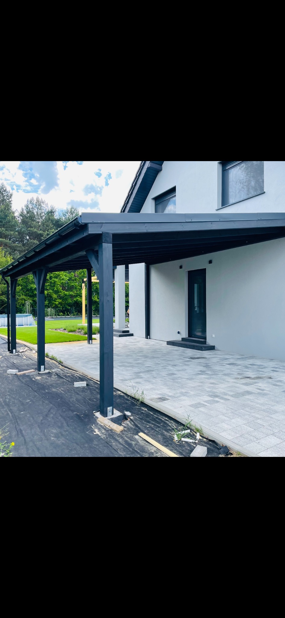 Nowoczesna, czarna konstrukcja zadaszenia przylegająca do jasnoszarego budynku z widoczną kostką brukową i niezabezpieczonym fragmentem gruntu z tyłu domu.