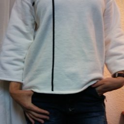 Zakład Krawiecki - Osoba w białej bluzie z czarnymi przeszyciami w kształcie krzyża i dżinsach z rękami w kieszeniach, na tle ściany i zasłony.