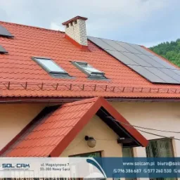 Czerwony dach domu jednorodzinnego z panelami fotowoltaicznymi, dwoma oknami dachowymi i kominem z cegły. W tle zielone wzgórza.