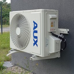 FIRMA KLIPO Stanisław Koba - Biała jednostka zewnętrzna klimatyzacji AUX zamontowana na szarej ścianie budynku z widocznymi przewodami. Widoczne mocowania i fragment trawnika.