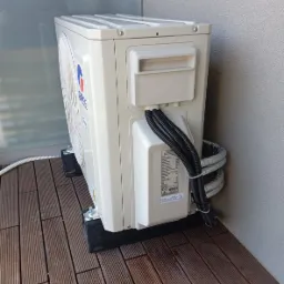 Boczny widok jednostki zewnętrznej klimatyzacji GREE na balkonie z drewnianą podłogą. Widoczne przewody w peszlu i podkładki antywibracyjne pod urządzeniem.