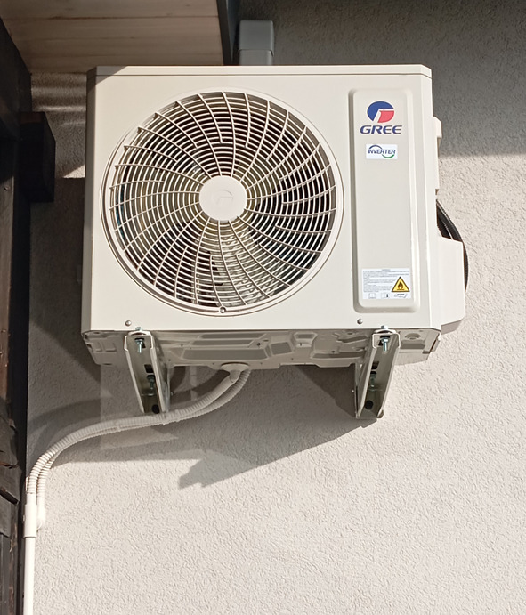 Biała jednostka zewnętrzna klimatyzatora marki Gree zamontowana na ścianie budynku za pomocą metalowych wsporników, widoczne przewody instalacyjne.