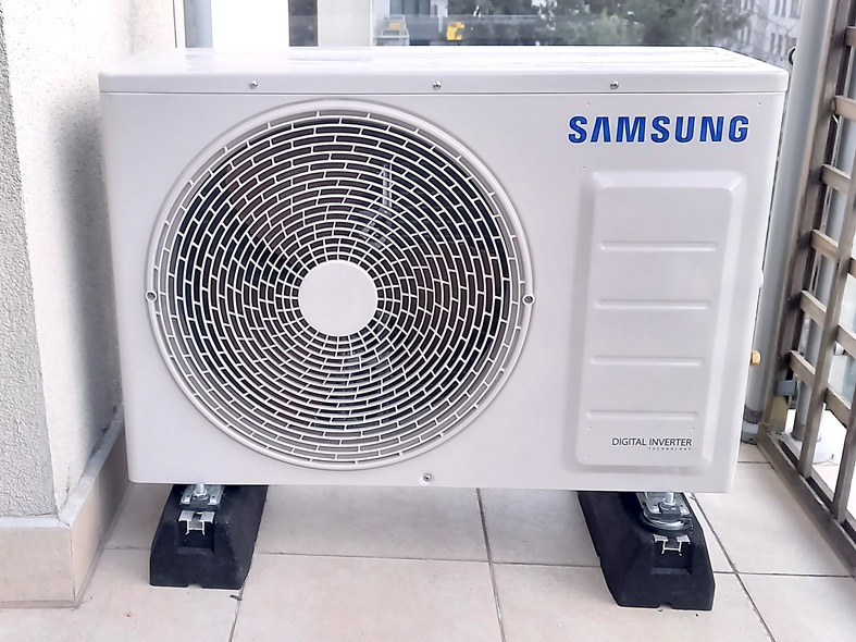 Zewnętrzna jednostka klimatyzacji Samsung Digital Inverter, zamontowana na czarnych podkładkach antywibracyjnych na balkonie z widoczną balustradą.