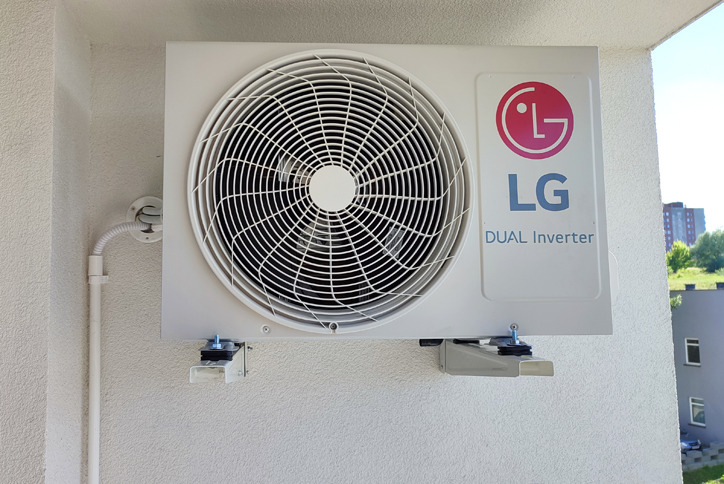 Zewnętrzna jednostka klimatyzatora LG Dual Inverter zamontowana na białej ścianie budynku, widoczne elementy montażowe i rurka odprowadzająca.