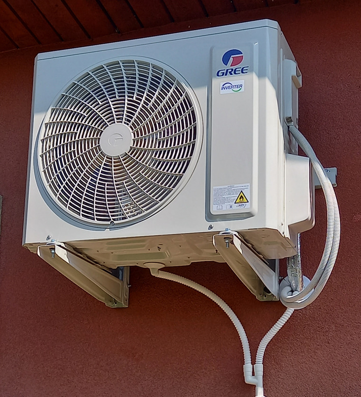 Zewnętrzna jednostka klimatyzacji marki Gree Inverter zamontowana na brązowej ścianie, widoczne białe rurki odprowadzające skropliny.