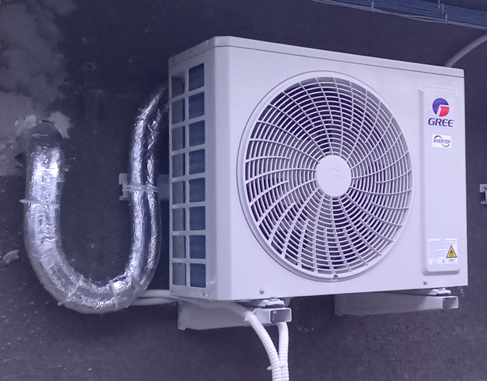 Zewnętrzna jednostka klimatyzacji marki Gree zamontowana na szarej ścianie, z widocznymi rurami w srebrnej izolacji i białymi kablami.