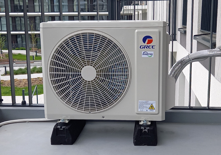 Zewnętrzna jednostka klimatyzacji Gree Inverter zamontowana na balkonie nowoczesnego budynku mieszkalnego, widoczne czarne podkładki antywibracyjne i rury odprowadzające.