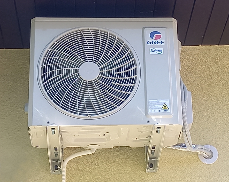 Zewnętrzna jednostka klimatyzatora marki Gree zamontowana na ścianie budynku z widocznymi elementami mocującymi i przewodami.