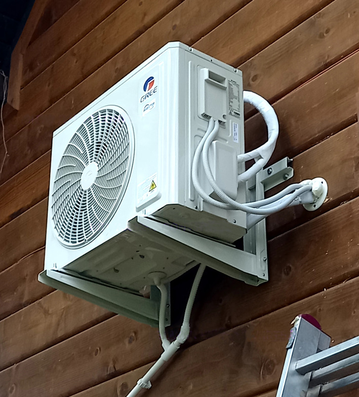 Biała jednostka zewnętrzna klimatyzacji marki Gree zamontowana na drewnianej elewacji, widoczne przewody w peszlach i drabina oparta o ścianę.