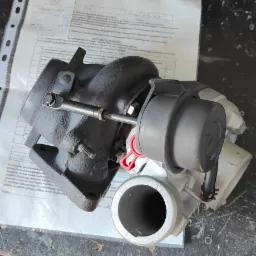 Turbosprężarka na stole warsztatowym z instrukcją montażu, widoczne ślady użytkowania na stole i częściowo widoczne buty osoby robiącej zdjęcie.