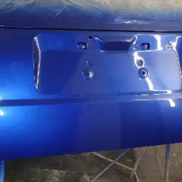 Błyszcząca, niebieska klapa bagażnika samochodu, świeżo po lakierowaniu, widoczne otwory montażowe na tablicę rejestracyjną i delikatne refleksy światła na gładkiej powierzchni.