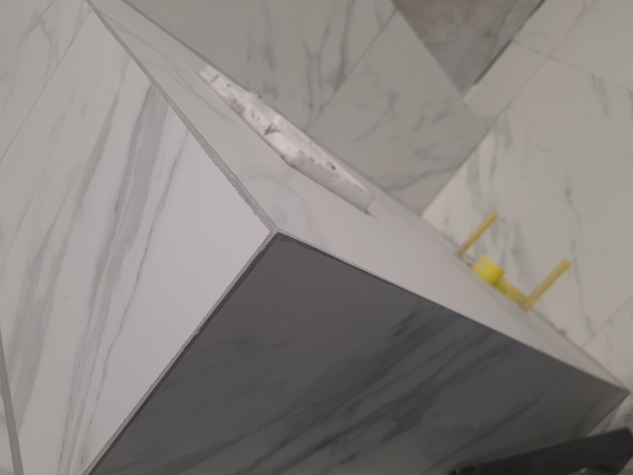 Szczegółowe ujęcie narożnika obudowy umywalki z płytek imitujących marmur, widoczne fugi i żółta poziomica, w tle płytki ścienne z tym samym wzorem.