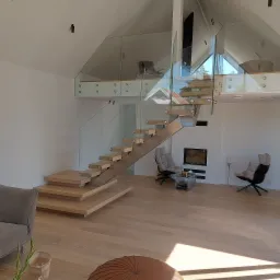 Nowoczesne schody z drewnianymi stopniami i szklaną balustradą w jasnym wnętrzu z wysokim sufitem i kominkiem. Minimalistyczny design.