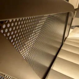 Nowoczesna, ażurowa barierka schodowa z metalu w kolorze beżowym, z widocznymi schodami w tle. Nietypowy kąt ujęcia podkreśla geometryczny wzór.