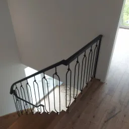 Kątowe ujęcie drewnianych schodów z czarną, metalową balustradą o dekoracyjnym wzorze. Widok z góry na drewniane stopnie i fragment podłogi z listwą przypodłogową.