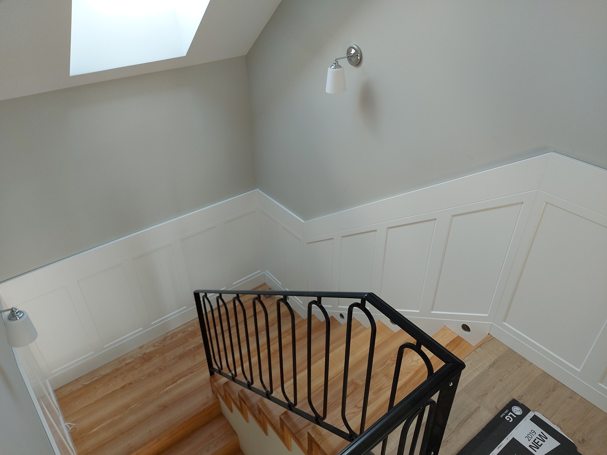 Klatka schodowa z drewnianymi stopniami, czarną metalową barierką i białymi panelami ściennymi. Widoczny fragment sufitu z oknem dachowym i lampa ścienna.