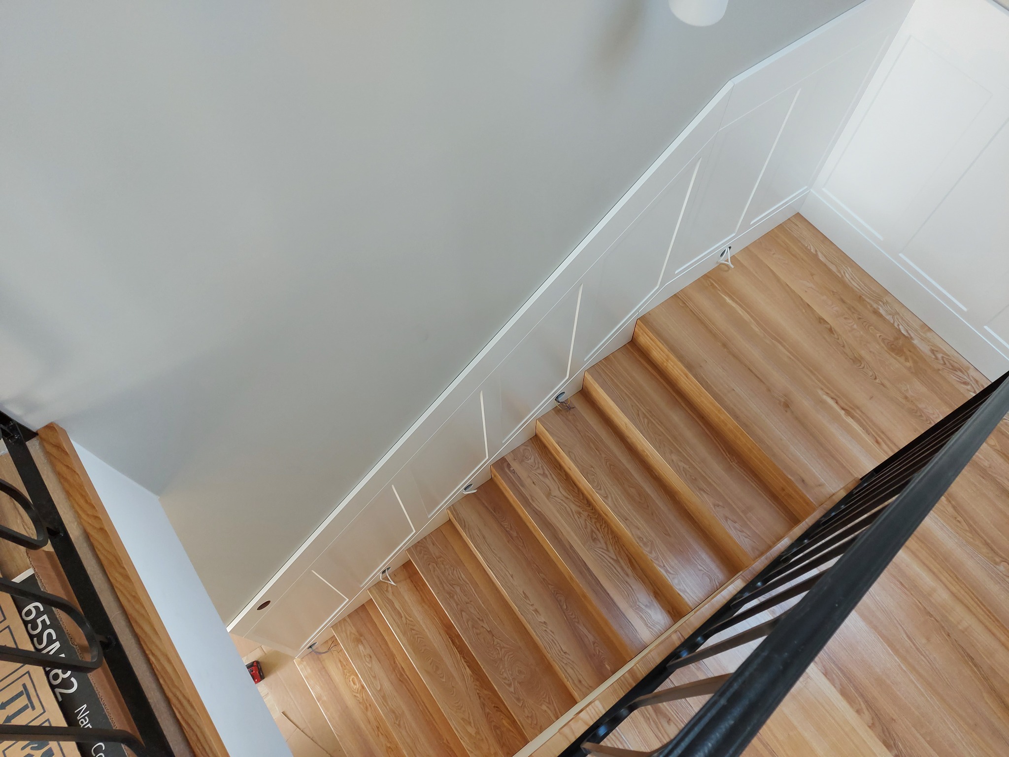 Drewniane schody z czarną, kutą balustradą, widziane z góry. Jasne drewno stopni kontrastuje z ciemną balustradą. Minimalistyczne białe ściany i listwy.