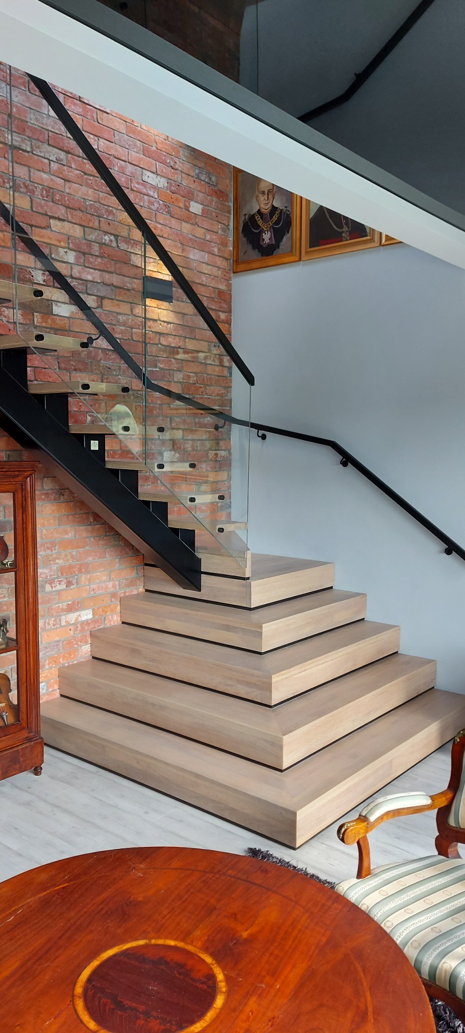 Nowoczesne schody z drewnianymi stopniami i czarnymi elementami metalowymi oraz szklaną balustradą, na tle ceglanej ściany i klasycznych mebli.