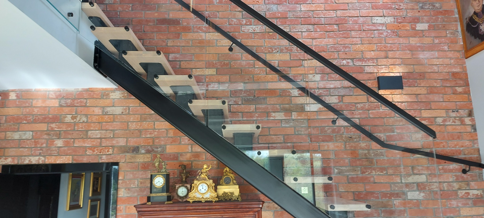 Nowoczesne schody z drewnianymi stopniami i szklaną balustradą na tle ściany z czerwonej cegły, z widocznymi zegarami na komodzie. Minimalistyczny design.