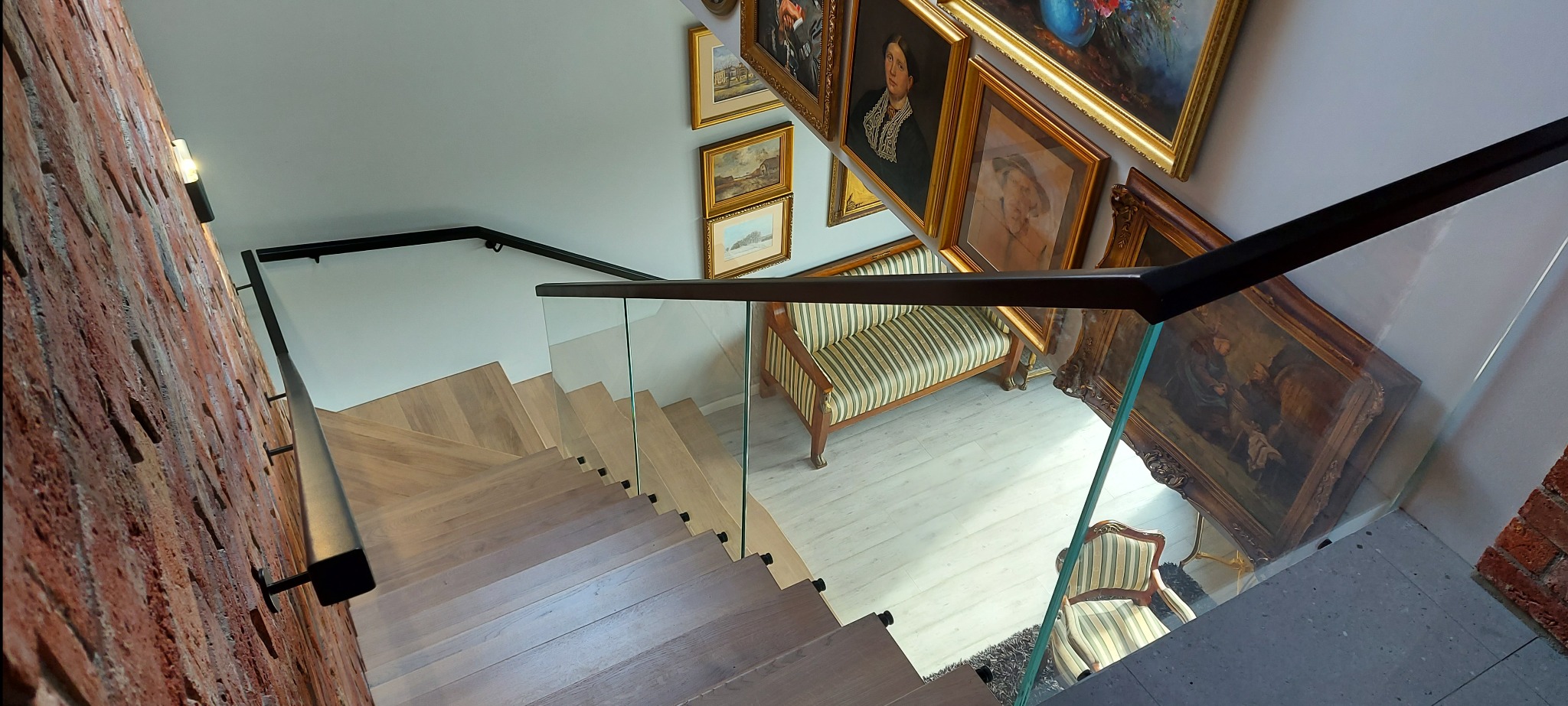 Nowoczesne schody z drewnianymi stopniami i szklaną balustradą, czarne poręcze, ceglana ściana, obrazy w złotych ramach, widok z góry.