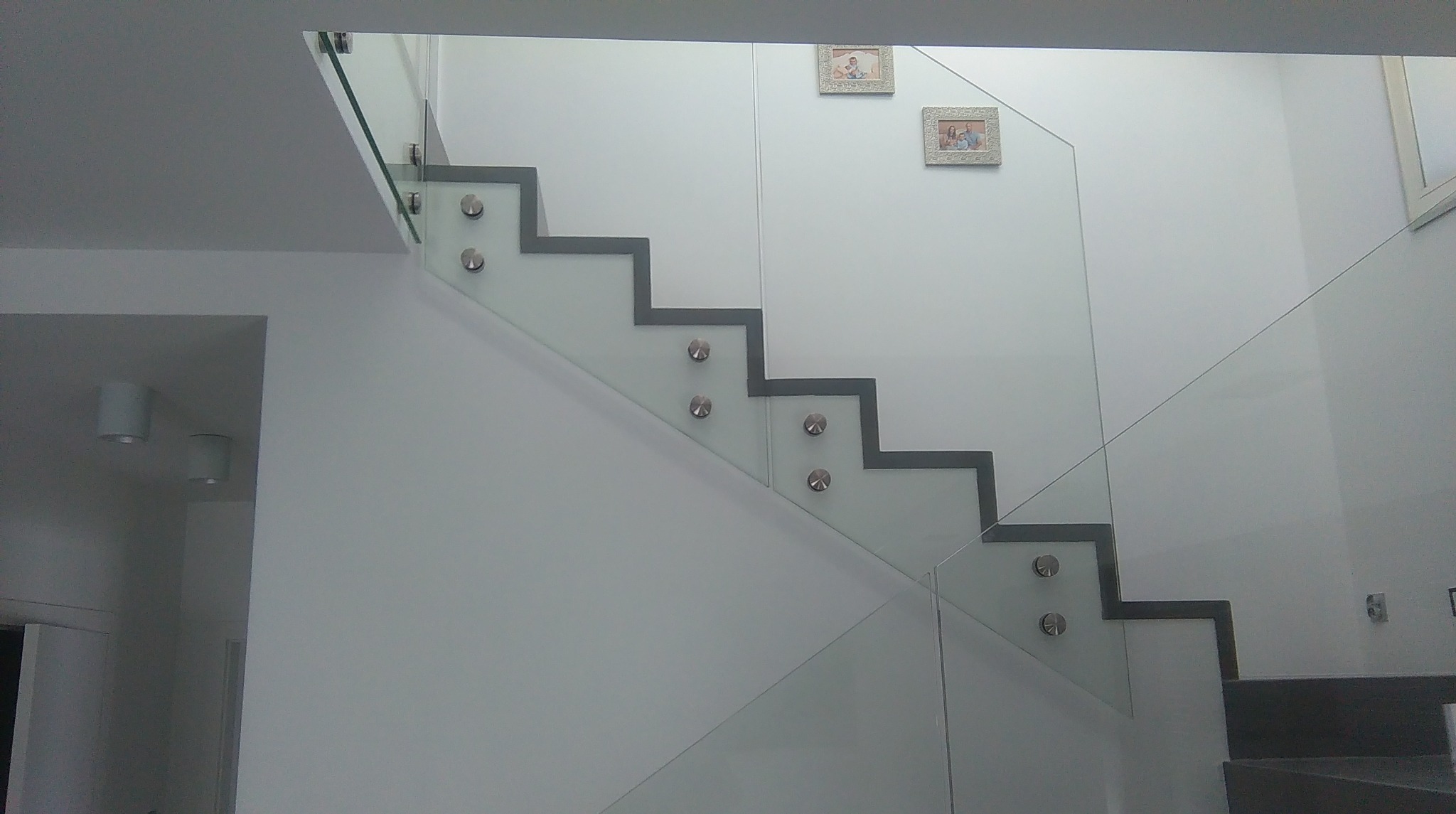 Minimalistyczne schody z szarymi stopniami i szklaną balustradą mocowaną na metalowych uchwytach. Na ścianie wiszą dwie małe ramki ze zdjęciami.